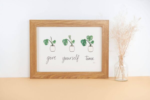 Framed print with hand lettered text 'give yourself time' and illustrations of a slowly uncurling monstera plant