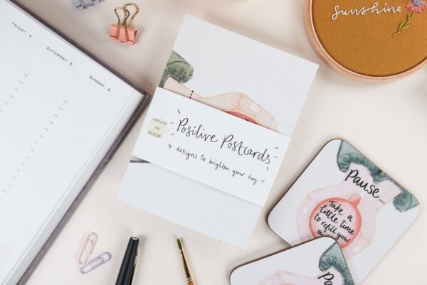 Positive postcard designs on a desk with matching coaster that reads 'Pause... take a little time to refill your cup'
