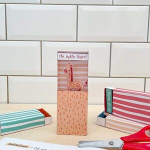 A completed matchbox craft kit stands with a surprise giraffe inside. Around the matchbox are craft kit paper and scissors.